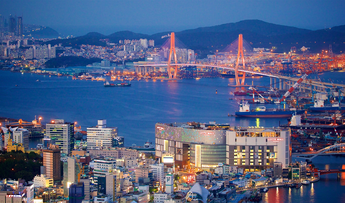 Foto panorâmica da cidade de Busan. Apresenta a cidade com muitos prédios, uma ponte grande sobre um rio e as luzes de prédios e da rua iluminando a cidade.